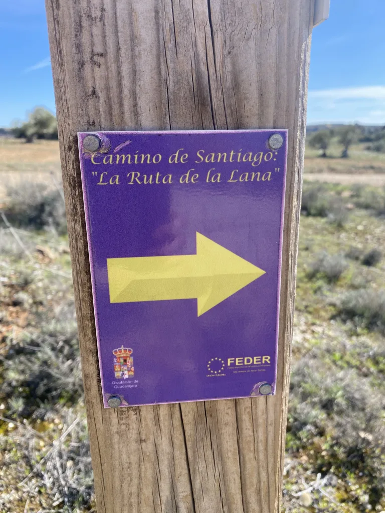 Señalización de madera en la Ruta de la Lana del Camino de Santiago, con flecha amarilla indicando la dirección.