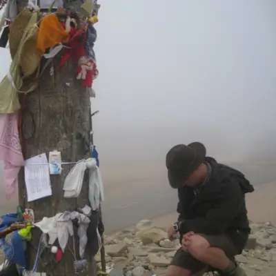 Peregrino arrodillado junto a la Cruz de Ferro en el Camino de Santiago rodeado de recuerdos y ofrendas, con niebla de fondo