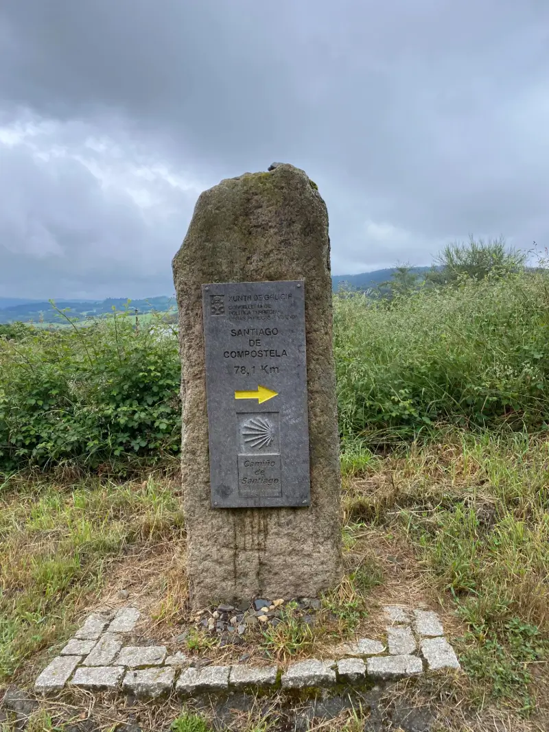 Mojón de piedra en el Camino de Santiago marcando 78,1 km hasta Compostela, rodeado de vegetación y con el cielo nublado.