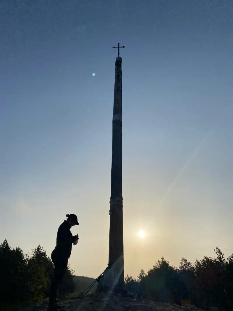 Silueta de un peregrino junto a una gran cruz de madera en el Camino de Santiago al amanecer, con el sol de fondo.