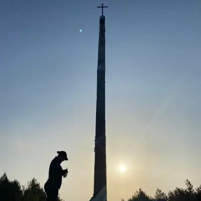 Peregrino rezando junto a la Cruz de Ferro en el Camino de Santiago al amanecer, con el sol de fondo y un cielo despejado.
