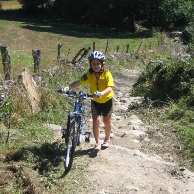 Niña con casco y equipo ciclista empujando su bicicleta por un sendero pedregoso del Camino de Santiago, rodeado de naturaleza.