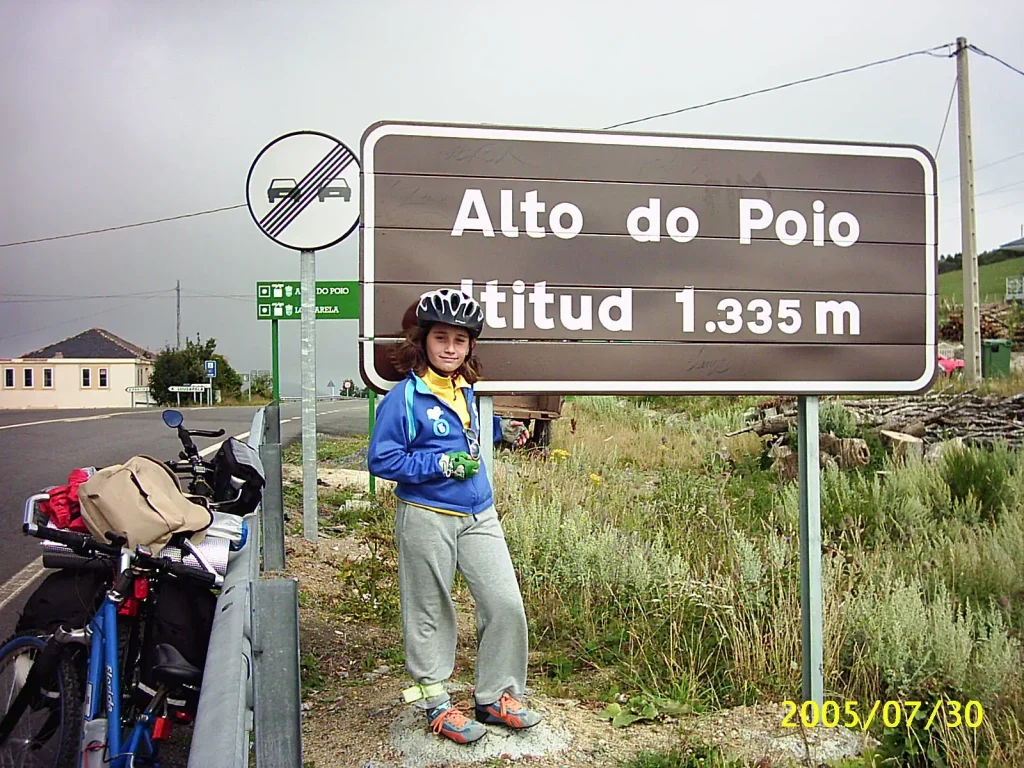 Niña ciclista con casco y chándal azul en el Alto do Poio, a 1.335 metros de altitud, junto a un cartel que marca la cima.