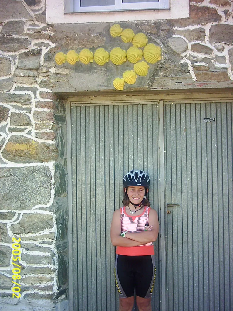 Niña con casco y ropa deportiva sonriendo frente a una pared con flechas de conchas amarillas, símbolo del Camino de Santiago.