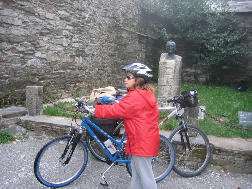 Niña con casco y chaqueta roja apoyada en su bicicleta junto a un busto de piedra en una parada del Camino de Santiago.