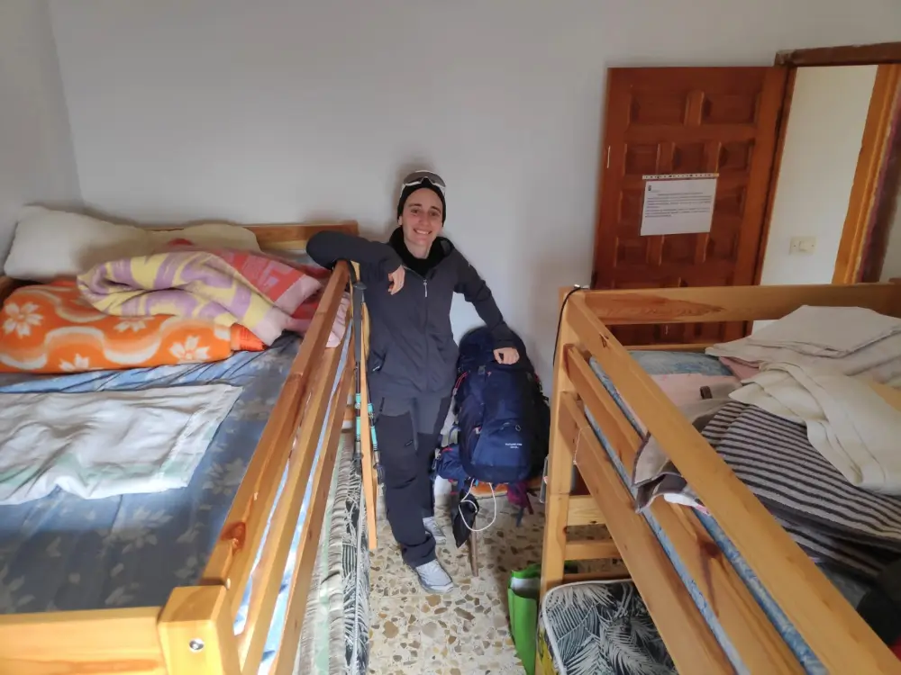 Mujer peregrina en un albergue del Camino de Santiago entre camas compartidas, con mochila, bastones y ropa de invierno