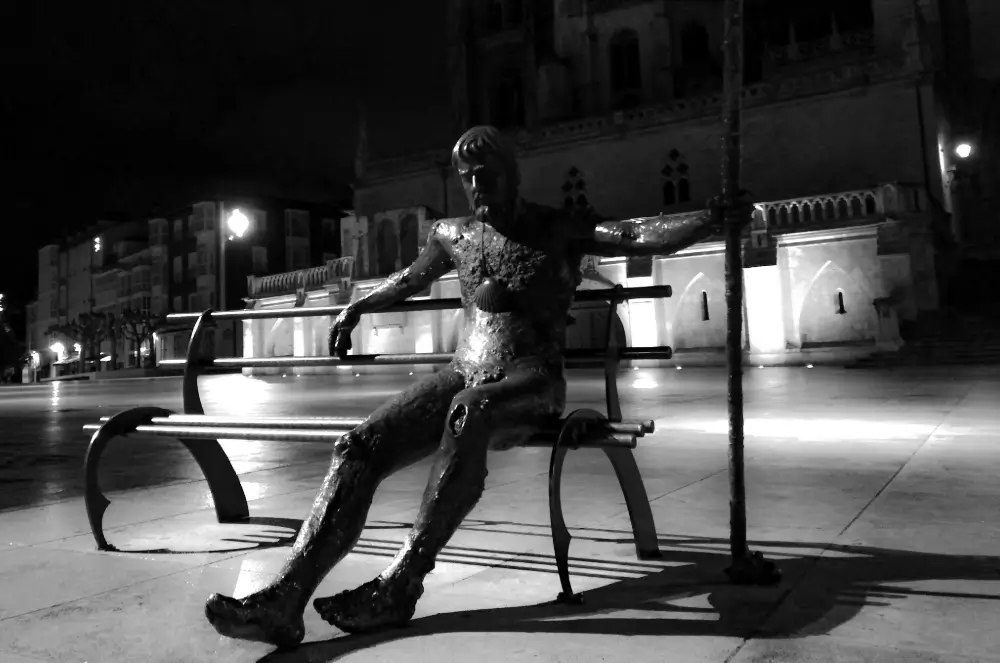 Escultura en bronce de un peregrino descansando en un banco de León, iluminada de noche, representando la fatiga y reflexión que acompañan a quienes hacen el Camino de Santiago.