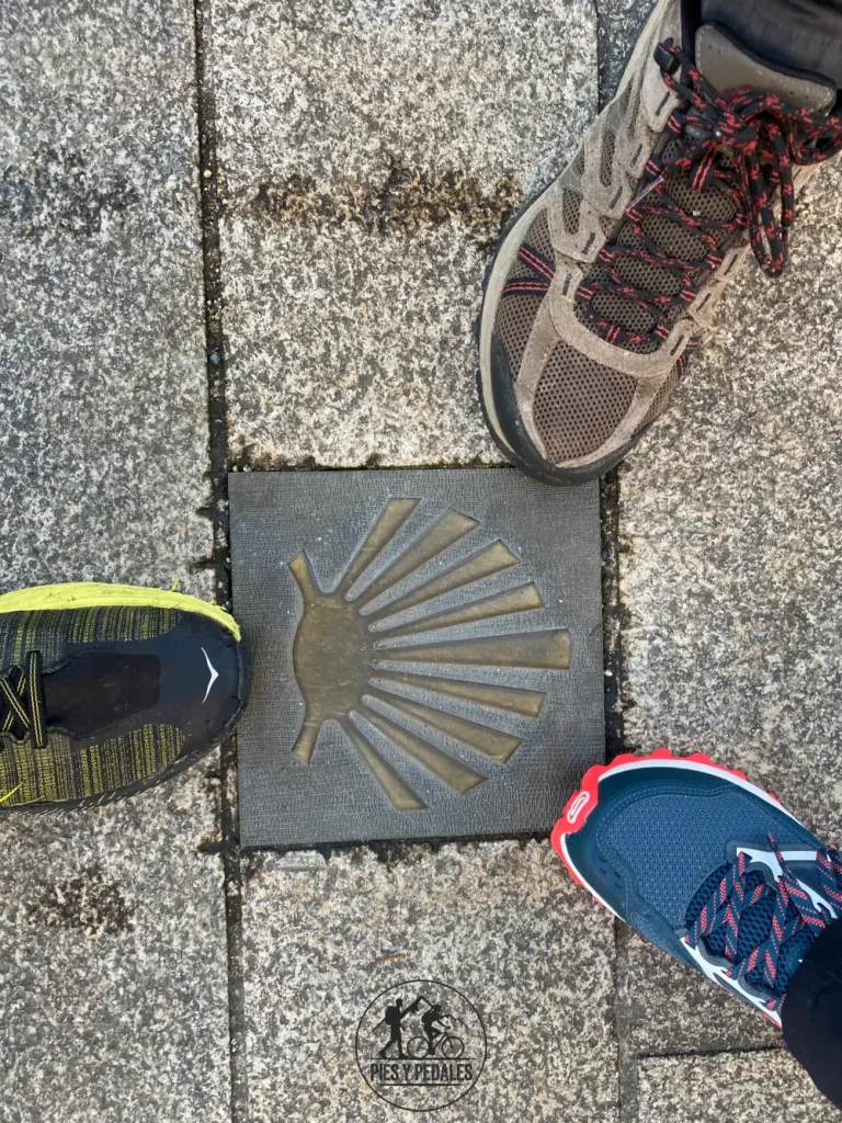 Zapatillas de tres peregrinos junto a una concha del Camino de Santiago en el suelo de piedra