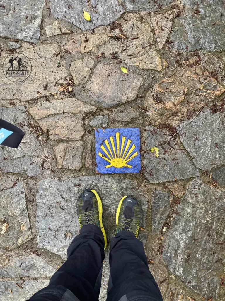 Pies de peregrino con zapatillas de trail sobre una losa con la flecha amarilla del Camino